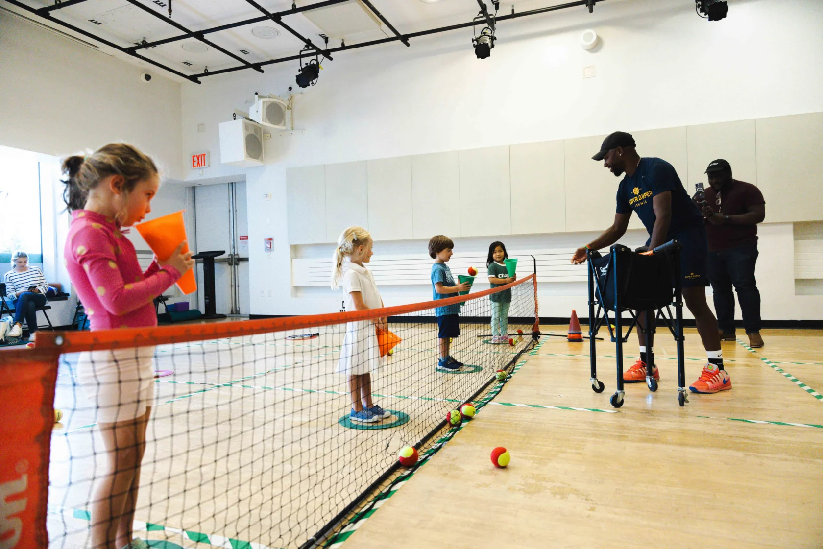 Children participate in an indoor tennis lesson, standing on one side of a net. An instructor assists them using a walker. Tennis balls are scattered on the floor.