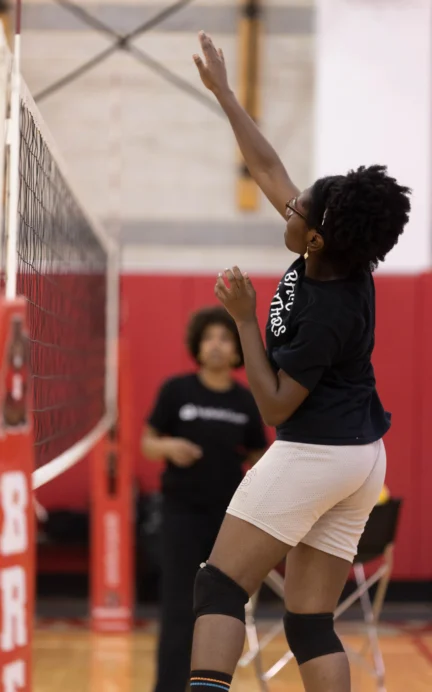 Person in black shirt and white shorts jumping to reach a volleyball net indoors, with another person in the background.