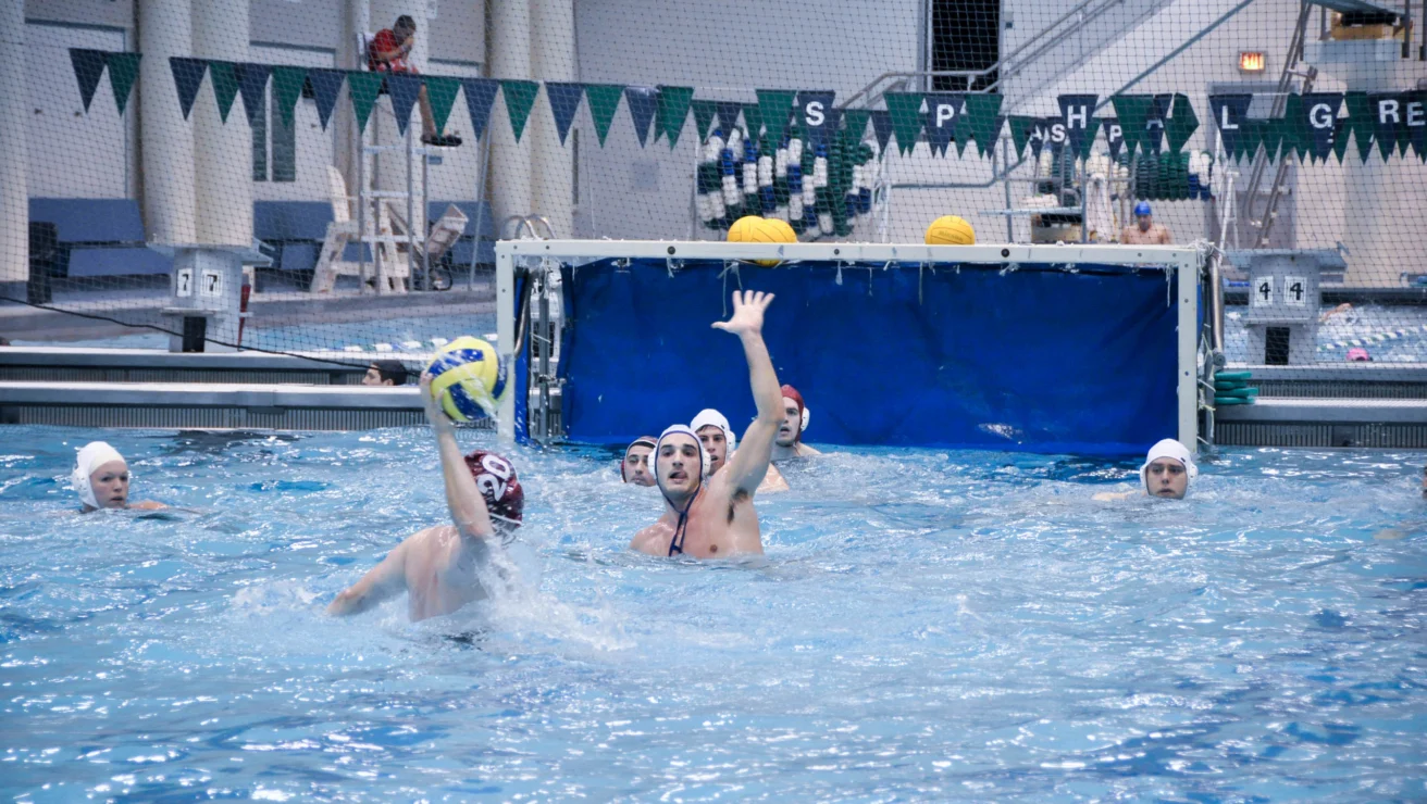 Water polo match in progress with players in white caps, one attempting a shot at goal while others defend. Blue and white pool setting with a goal and pennant flags in the background.