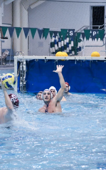 Water polo match in progress with players in white caps, one attempting a shot at goal while others defend. Blue and white pool setting with a goal and pennant flags in the background.