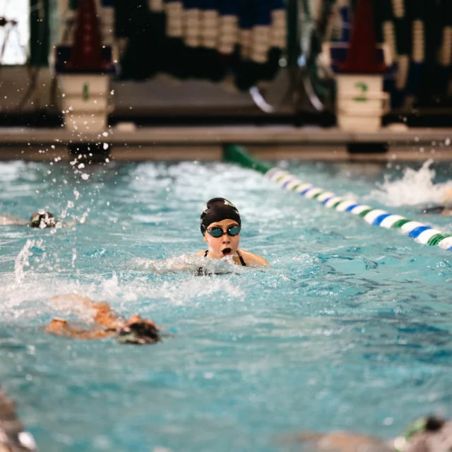 A swimmer in a black cap and goggles swims a breaststroke in a pool, surrounded by lane markers and splashing water.