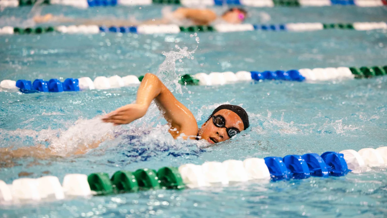 A swimmer wearing goggles competes in a freestyle race in a swimming pool, surrounded by blue, green, and white lane markers.