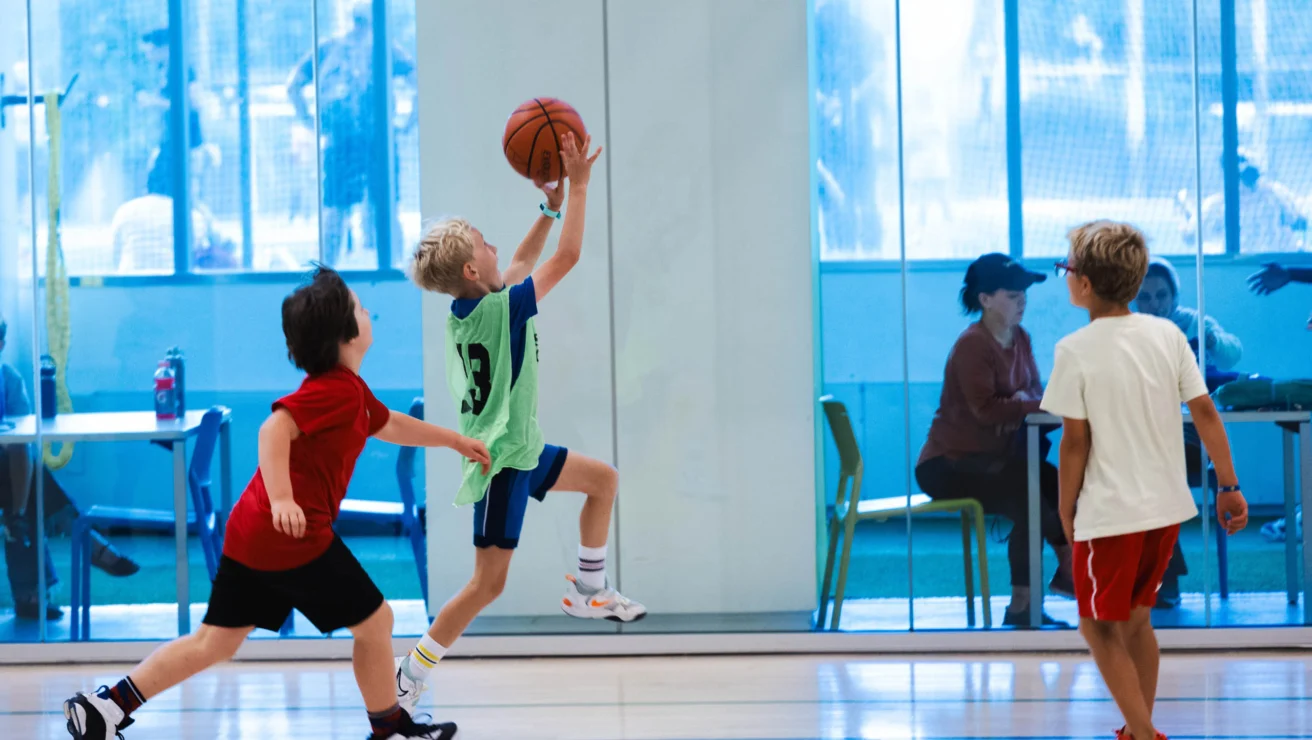 Three children play basketball indoors, with one jumping to shoot the ball.