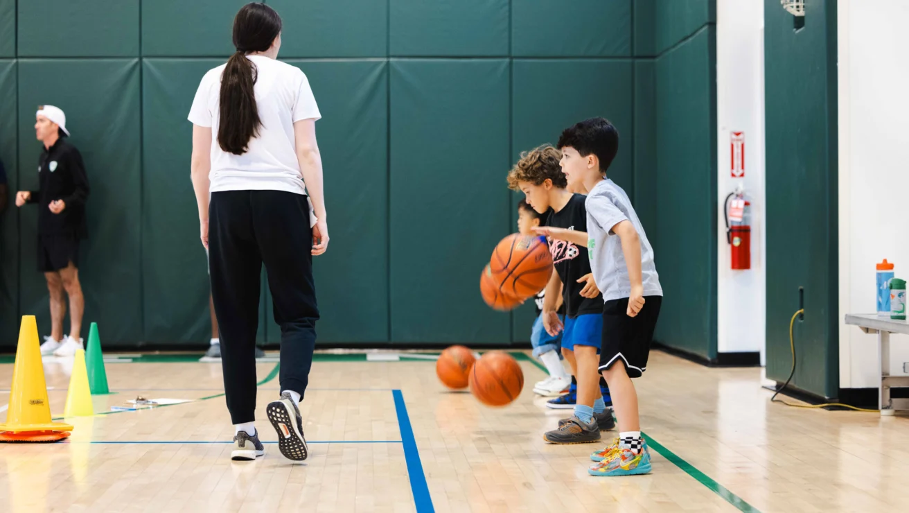 Children practice dribbling basketballs in a gym under an adult's supervision, with cones and hoops in the background.