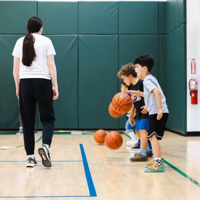 Children practice dribbling basketballs in a gym under an adult's supervision, with cones and hoops in the background.
