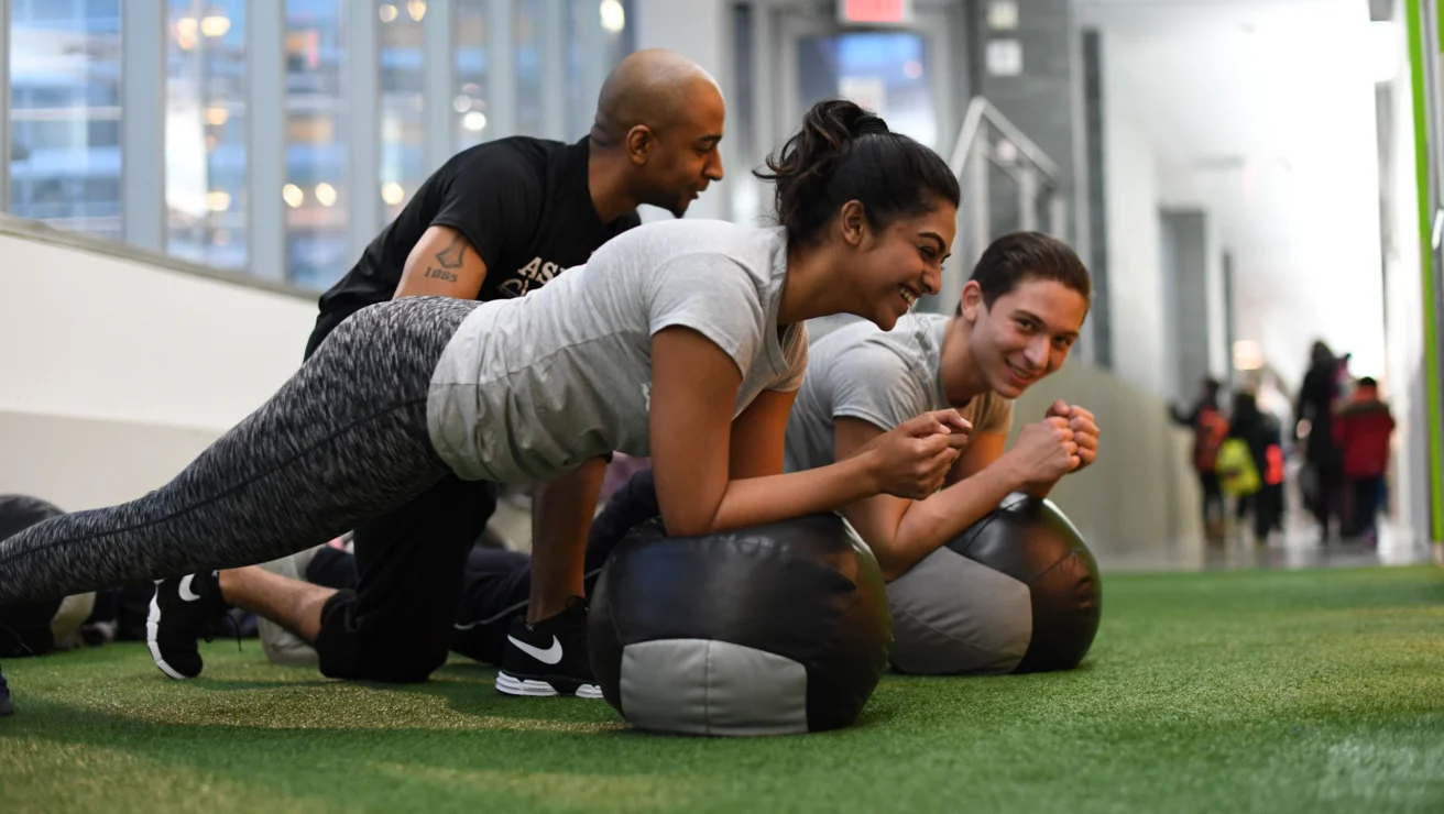 Three people in a gym perform planks on exercise balls, guided by an instructor. They are on a green indoor turf with large windows in the background.