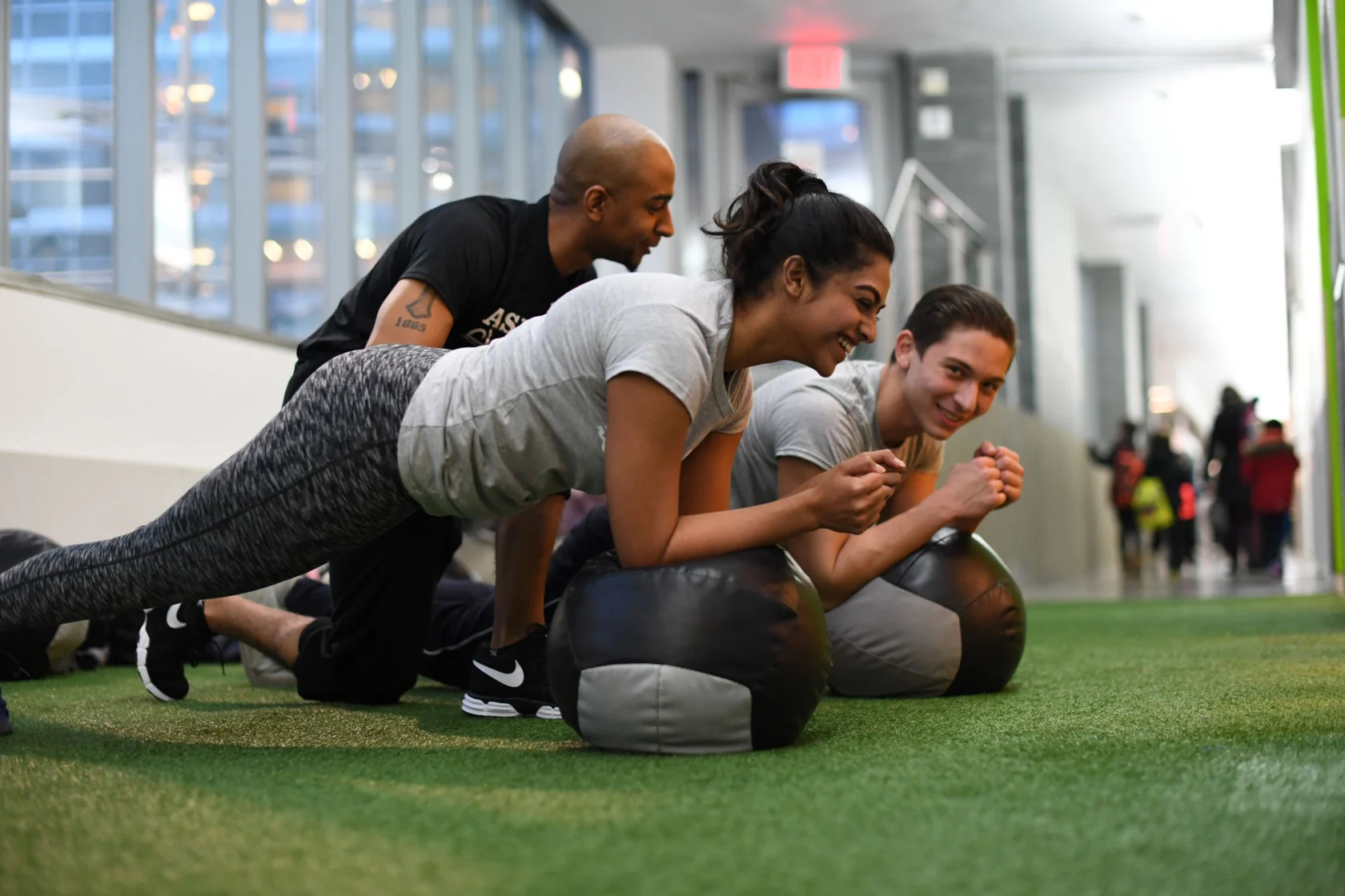 Three people in a gym perform planks on exercise balls, guided by an instructor. They are on a green indoor turf with large windows in the background.