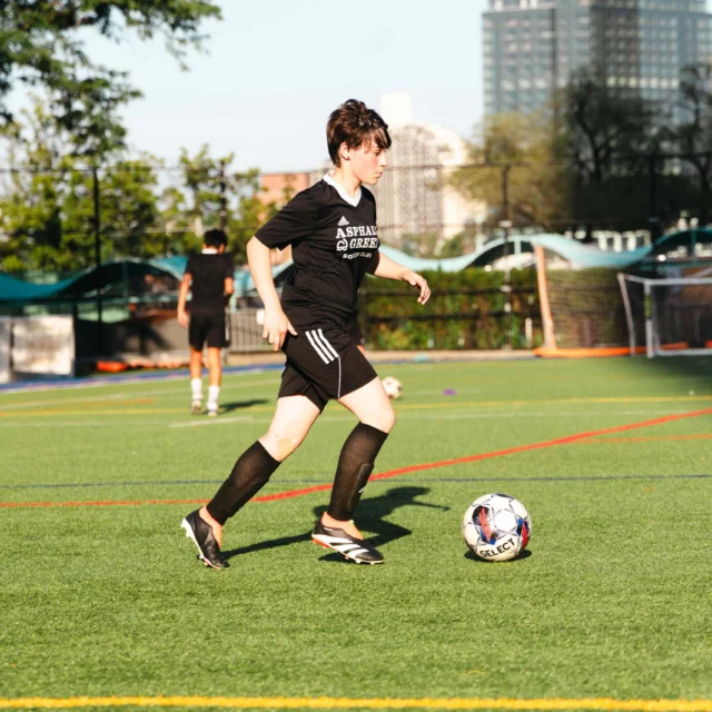 A person in a black soccer uniform is dribbling a ball on a green soccer field. Trees and buildings are visible in the background.