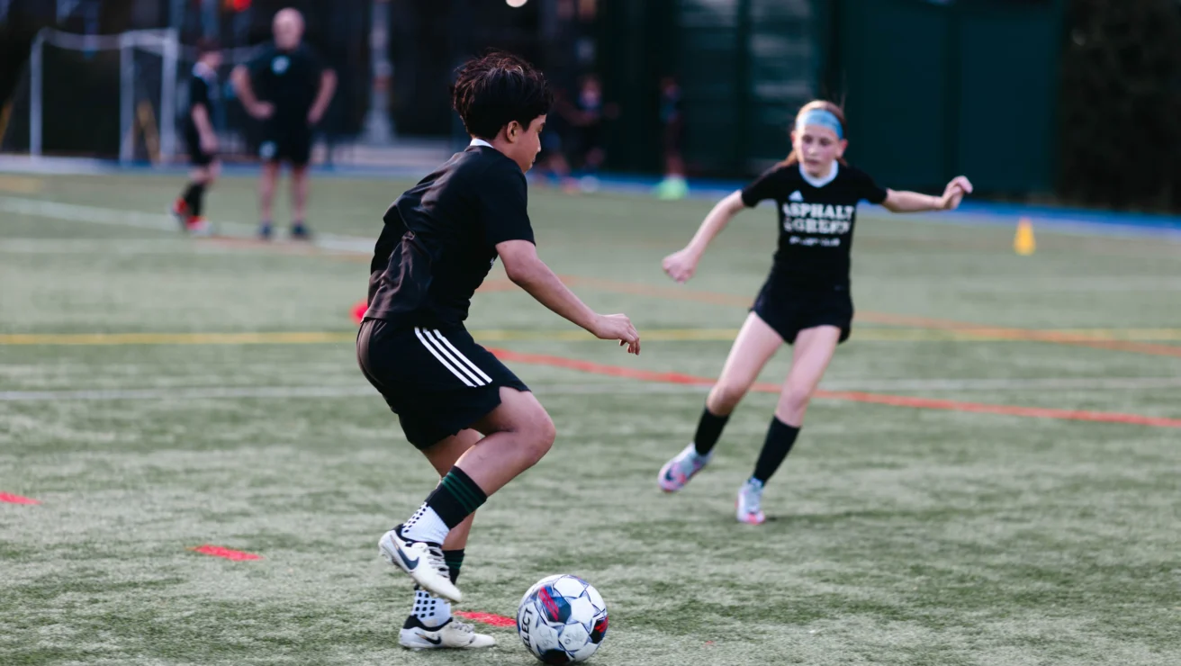 Two young soccer players in black outfits on a field; one is dribbling a ball while the other defends.