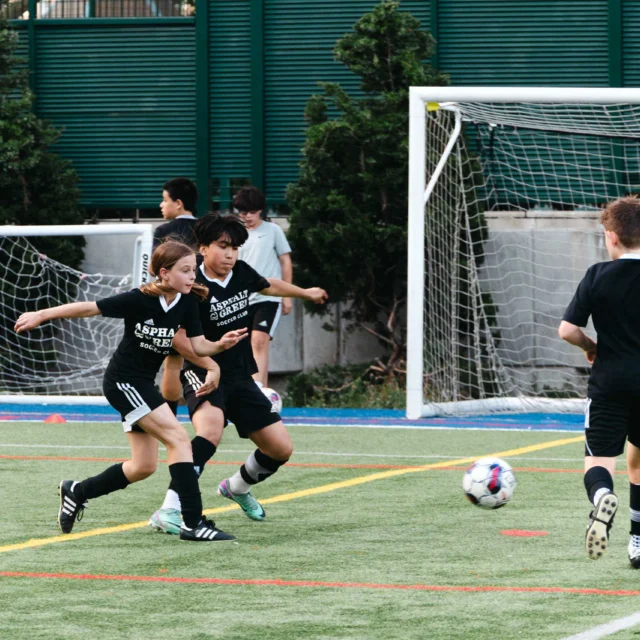Children playing soccer on a field, with some in black jerseys running and chasing the ball near a goalpost. Trees and a fence are in the background.