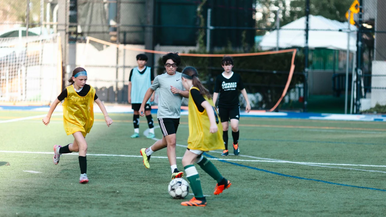 Children playing soccer on a field, with some wearing yellow pinnies and others in gray. They're actively pursuing the ball.