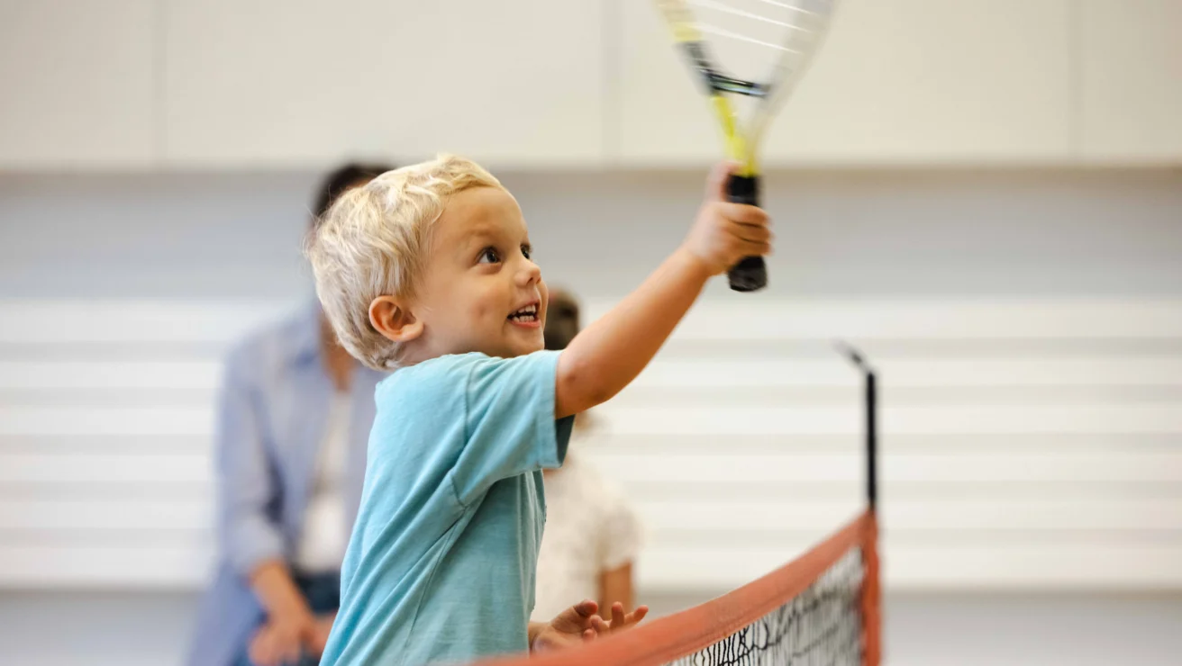 A child in a blue shirt joyfully swings a tennis racket near a net indoors, with a blurred person in the background.