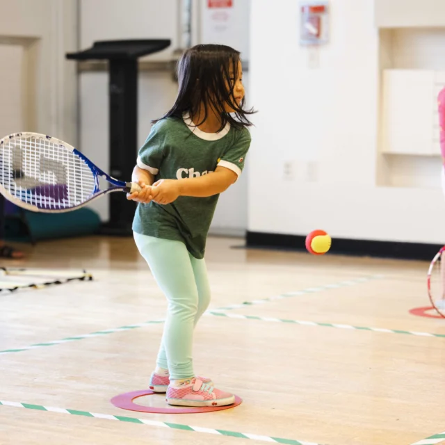 A young girl in a green shirt and teal leggings plays tennis indoors, swinging a racket at a colorful ball.