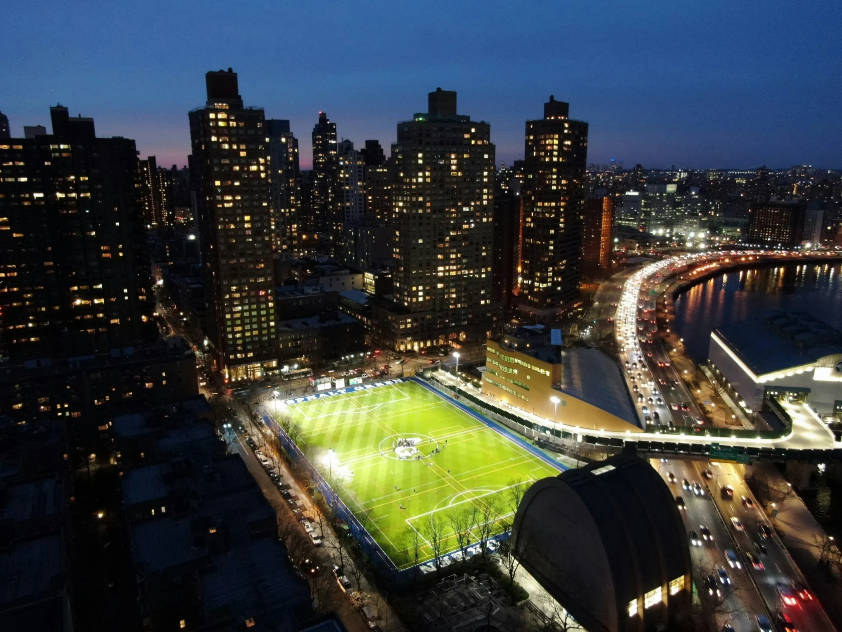 Aerial view of a brightly lit soccer field surrounded by tall buildings at night, with a highway and river on the right.