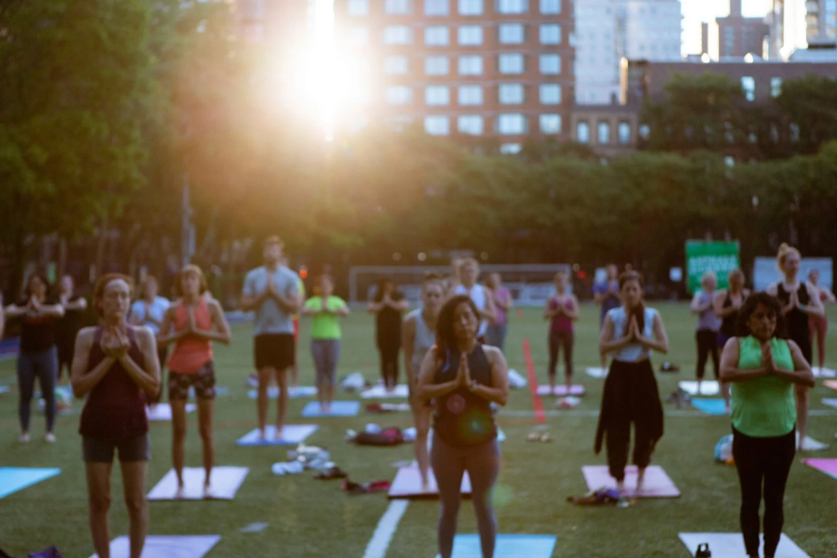 A group of people practicing yoga on mats in a park during sunset, with trees and buildings in the background.