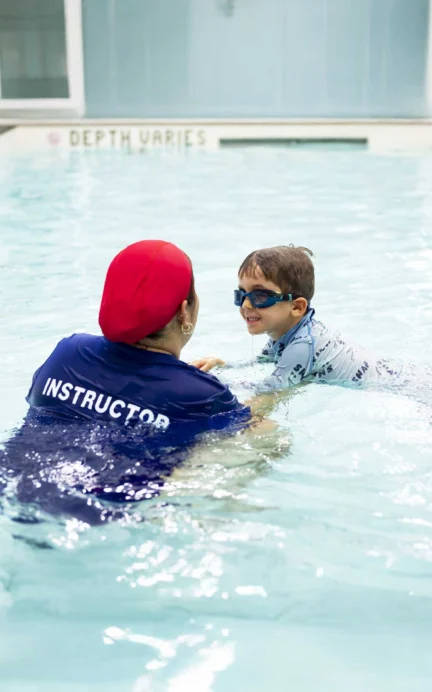 A swim instructor wearing a red head covering assists a child in blue goggles and clothing in a pool.
