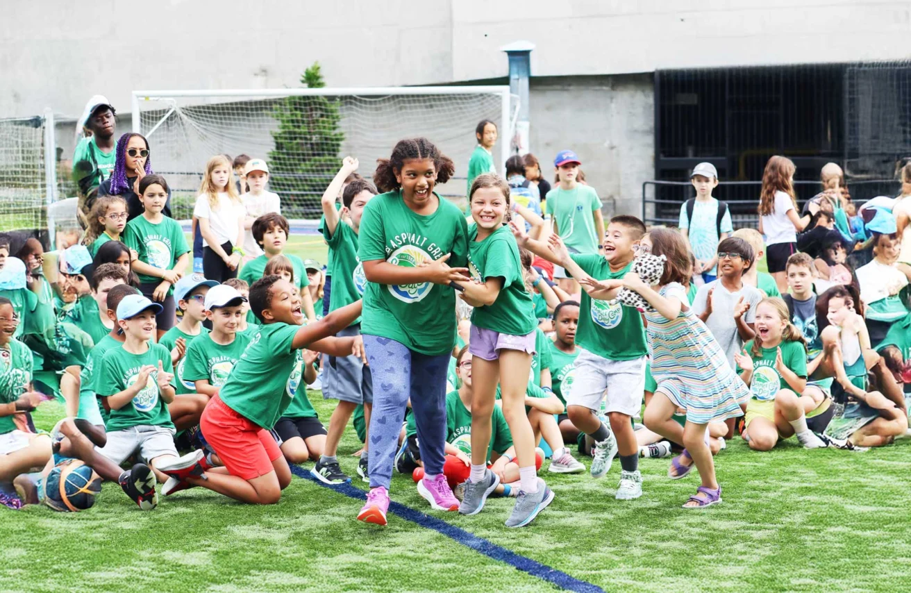 A group of children wearing green shirts are gathered outdoors on a sports field. Two girls in the center are holding hands, with others cheering and clapping around them.