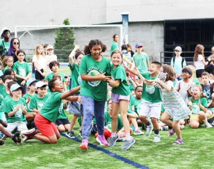 A group of children wearing green shirts are gathered outdoors on a sports field. Two girls in the center are holding hands, with others cheering and clapping around them.