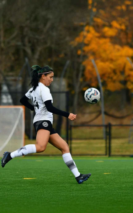 Soccer player in a white and black uniform runs with a soccer ball on a green field. Autumn trees and a goalpost are in the background.