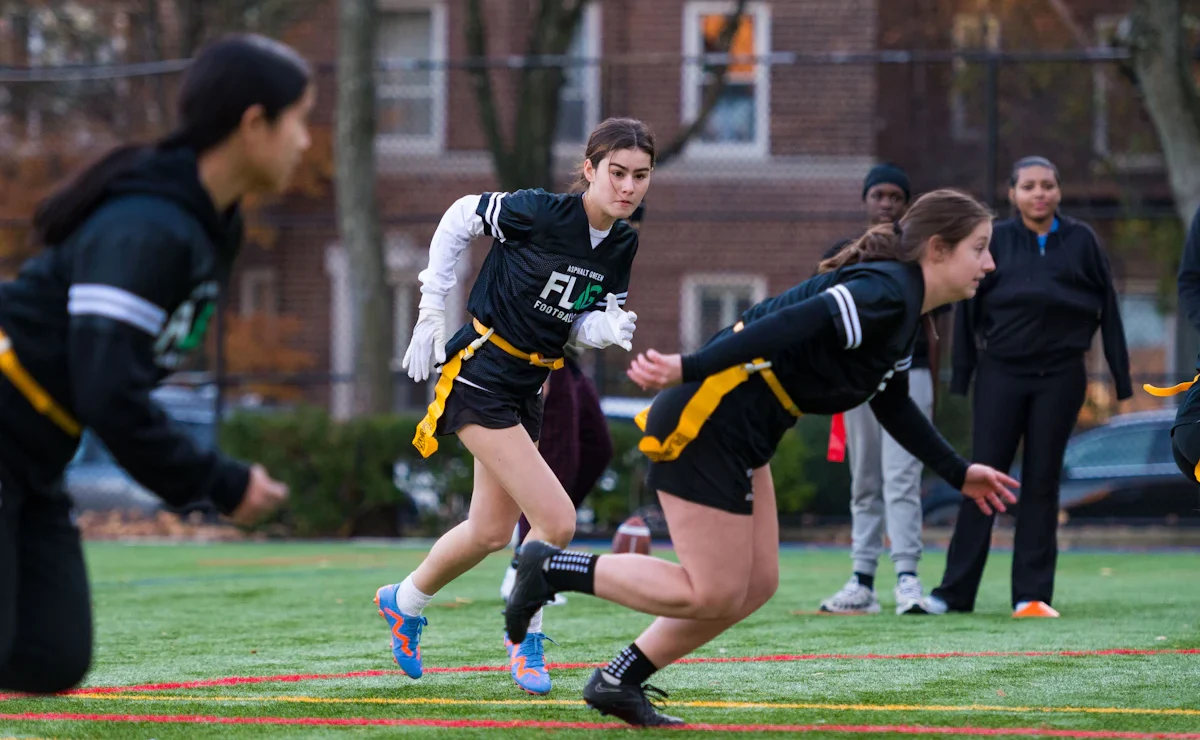 People playing flag football on a field; a focused player in the center runs while others are in motion. Brick buildings and trees are visible in the background.