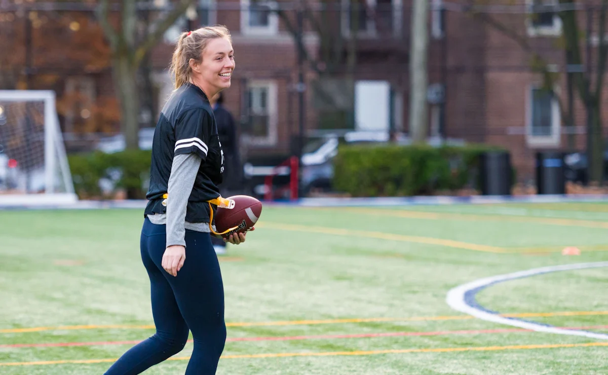 A woman in a black shirt and leggings is holding a football on a grassy field with buildings in the background.
