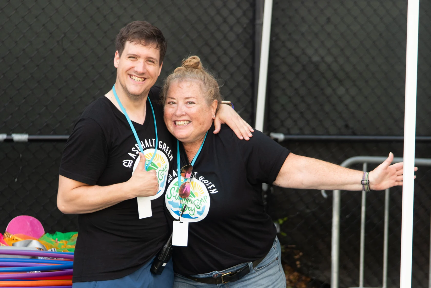 Two smiling people in matching black shirts with a colorful logo, standing side by side in front of a fence, one giving a thumbs-up.