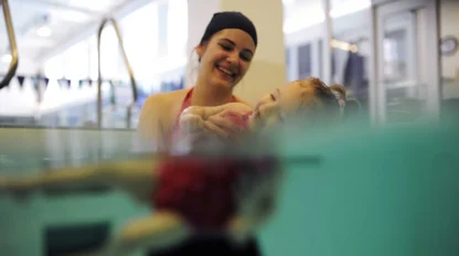 An adult and a child are in a swimming pool. The adult is smiling, holding the child, who is partially submerged in the water.