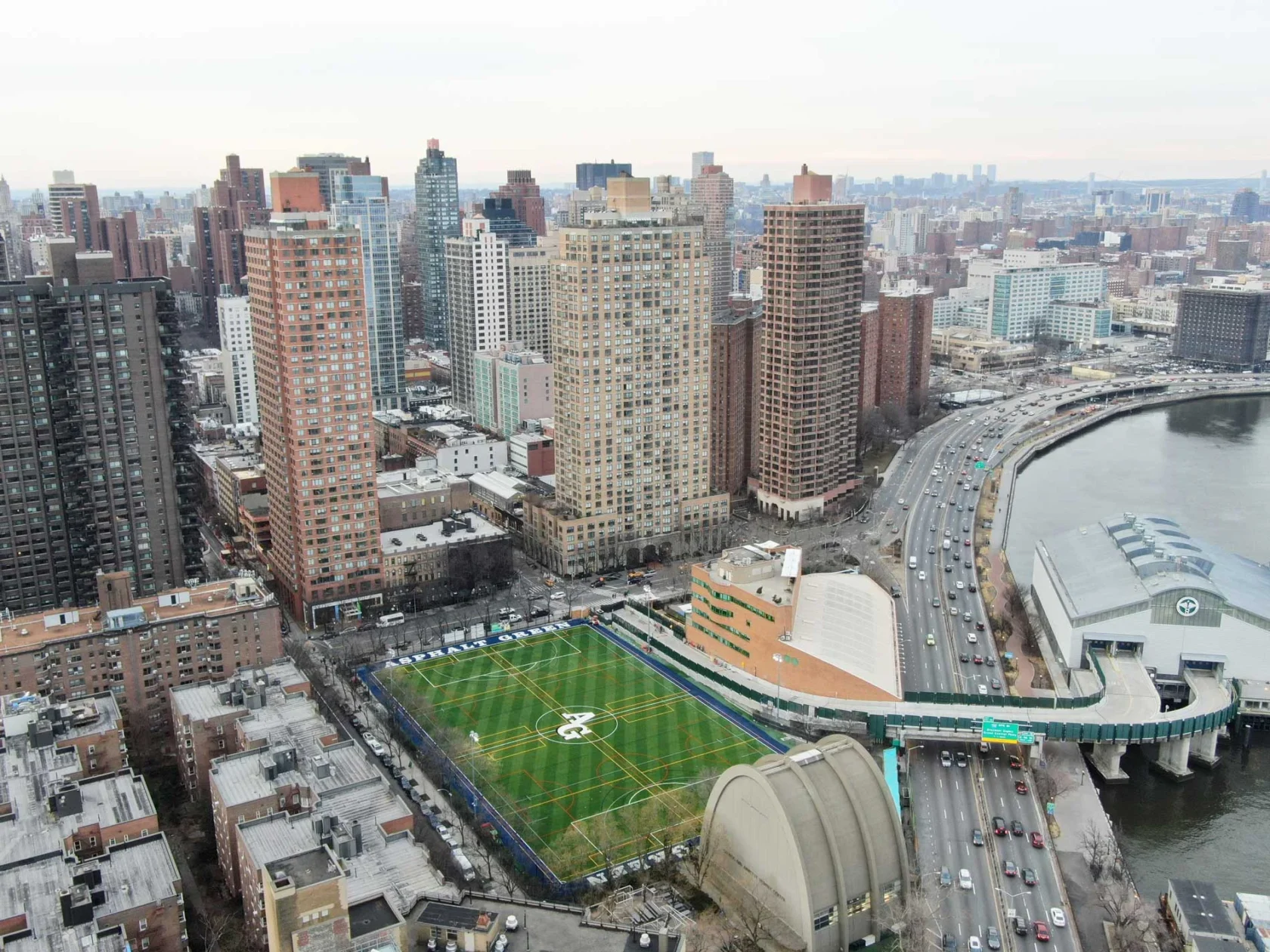 Aerial view of a cityscape with tall buildings, a sports field, and a curved riverside road.