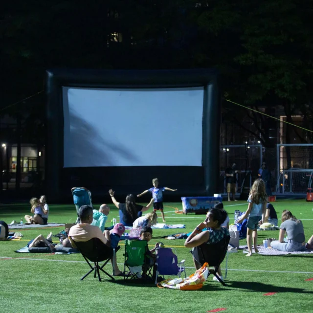 People sitting on blankets and chairs on a grassy field watching a large outdoor screen at night.