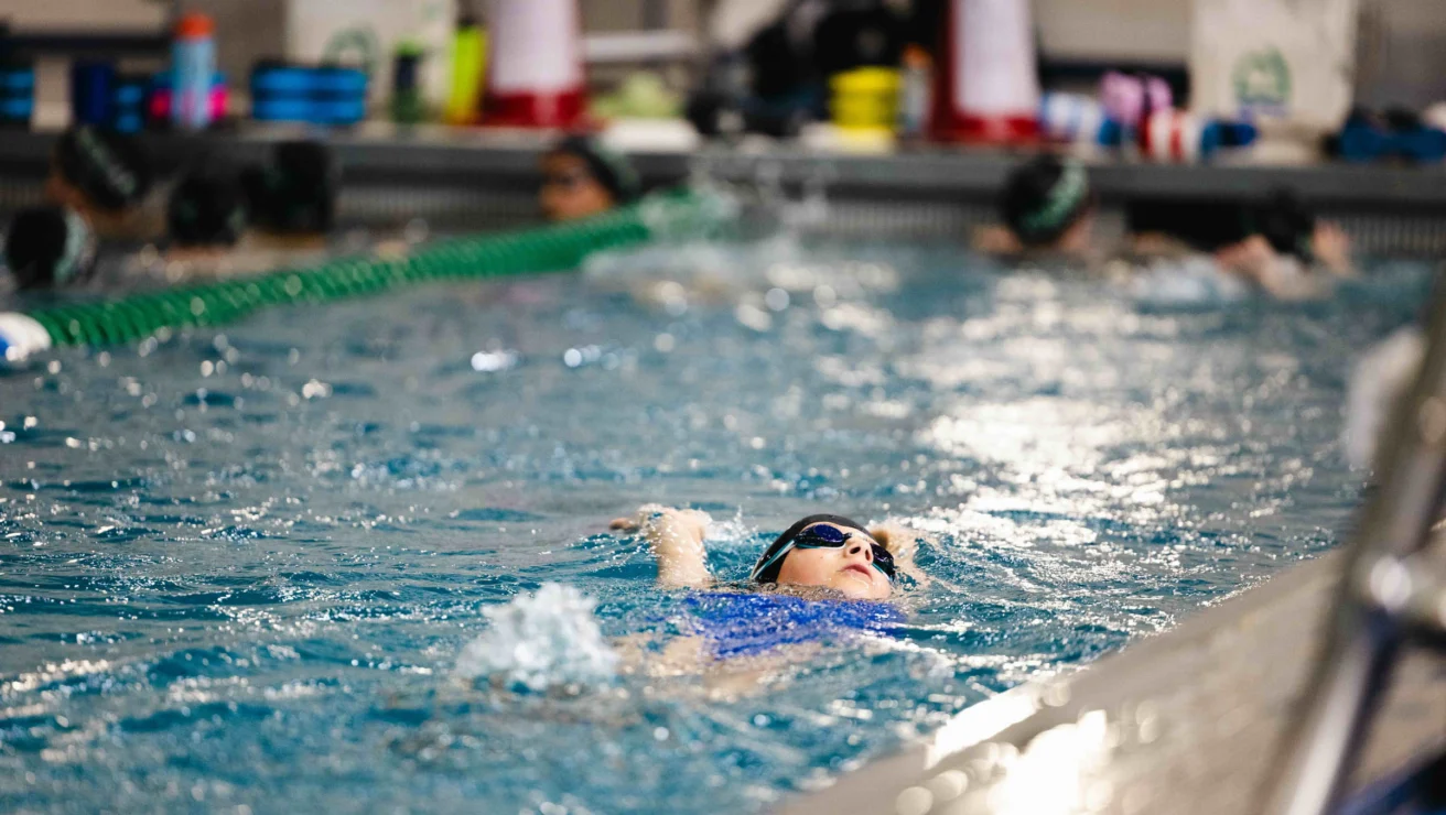 A swimmer in blue swimwear and goggles is doing the backstroke in an indoor pool. Other swimmers are in adjacent lanes.
