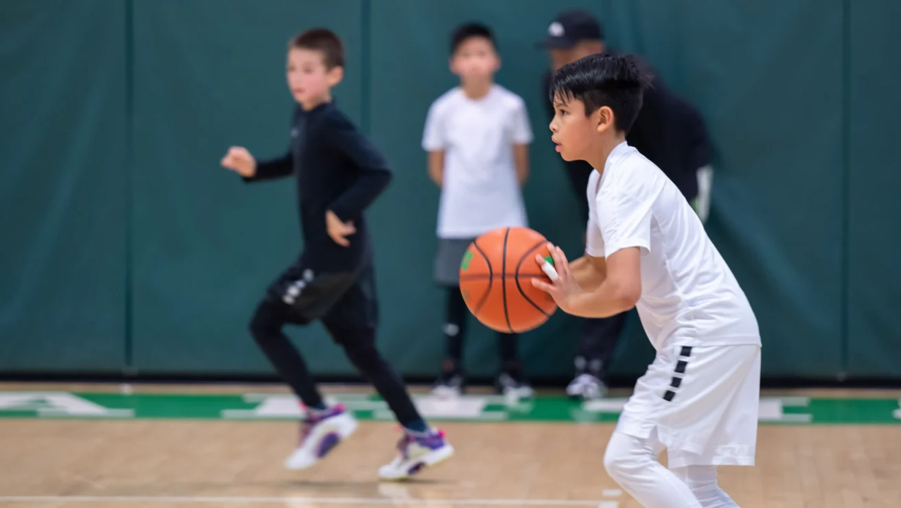 A young boy in a white jersey holds a basketball, preparing to pass, while other players and a coach are visible in the background on an indoor court.