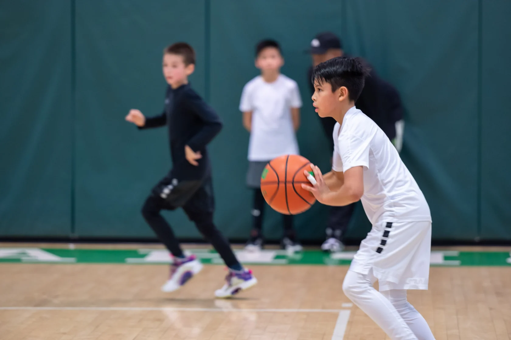 A young boy in a white jersey holds a basketball, preparing to pass, while other players and a coach are visible in the background on an indoor court.