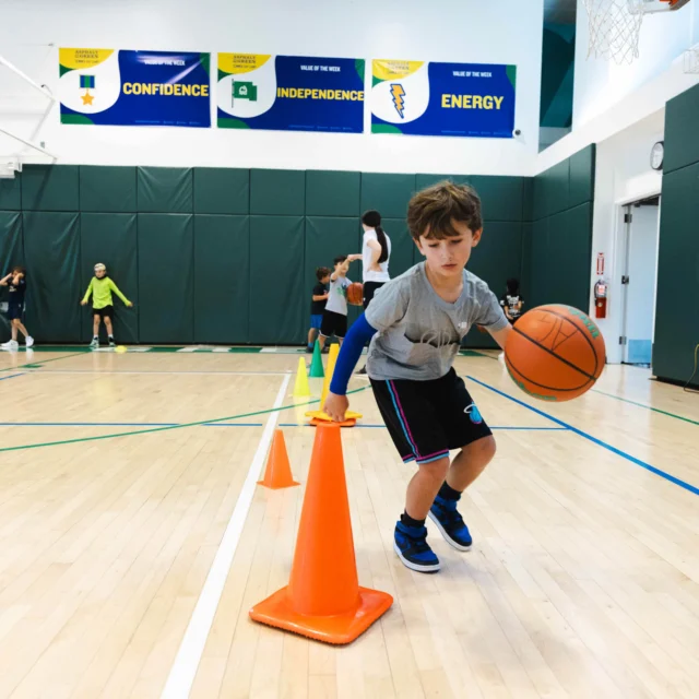 A child dribbles a basketball around orange cones in a gym. Other children are visible in the background, practicing various drills. Banners on the wall display "Confidence," "Independence," and "Energy.