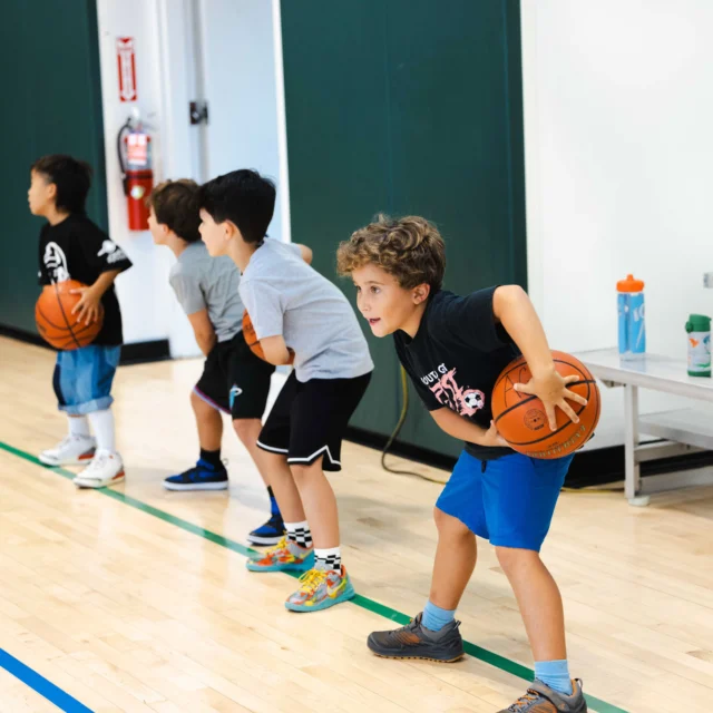 Four children lined up on a basketball court, each holding a basketball and preparing to dribble.