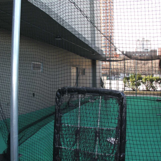 Indoor batting cage with nets and green flooring, surrounded by walls and a ceiling. A metal frame and sports equipment are visible.
