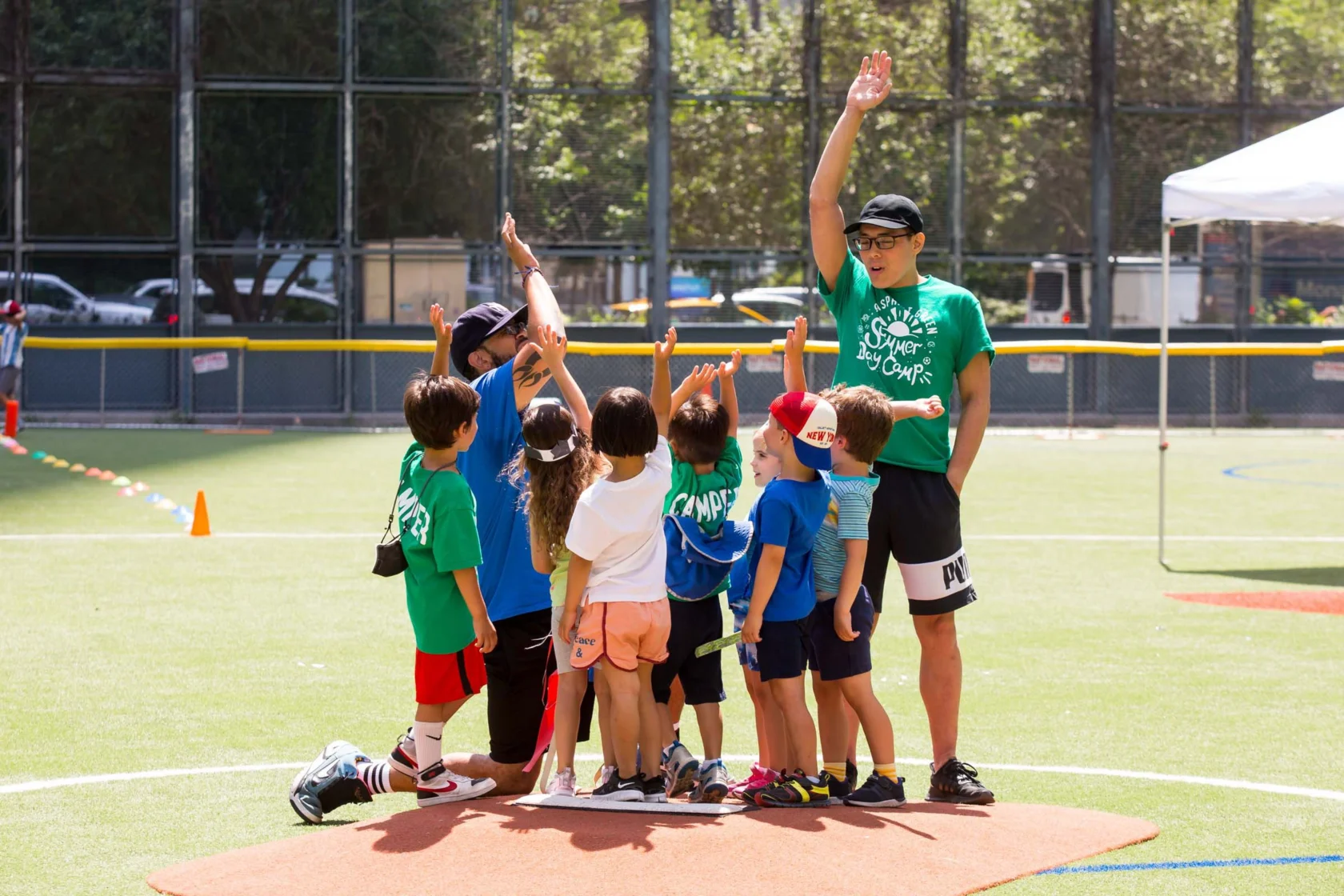 Children and two adults gather on a baseball field, raising hands in a group huddle. They wear casual sports attire, with shirts labeled "Camper" and "Instructor.