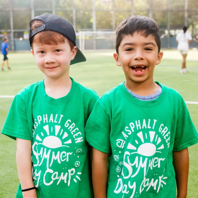Two boys in green "Asphalt Green Summer Day Camp" shirts stand on a sports field, smiling. Other children are playing in the background.