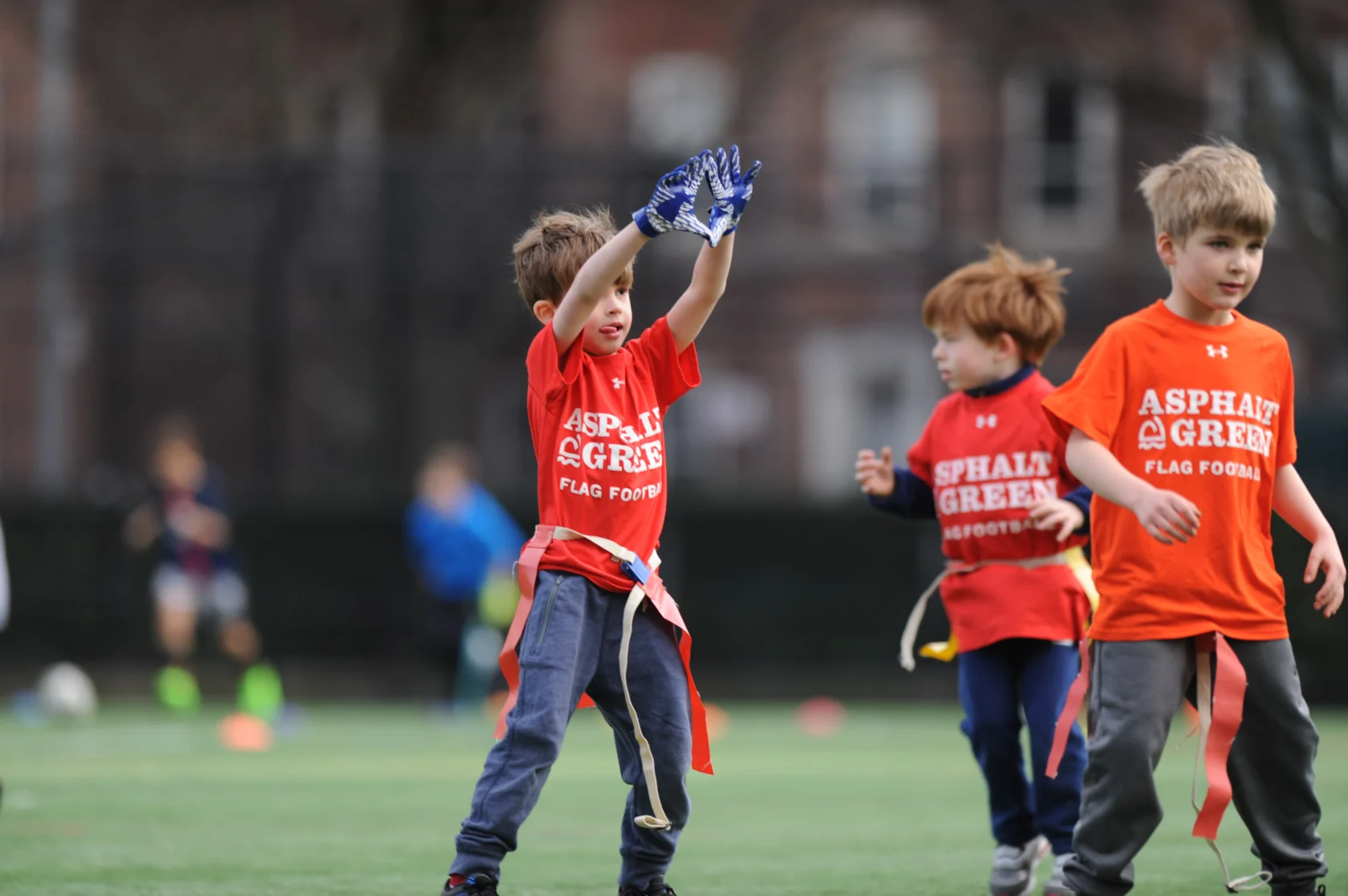Young children play flag football on a grassy field, wearing orange "Asphalt Green Flag Football" shirts and colorful flags. One child raises his arms, and two others run nearby.