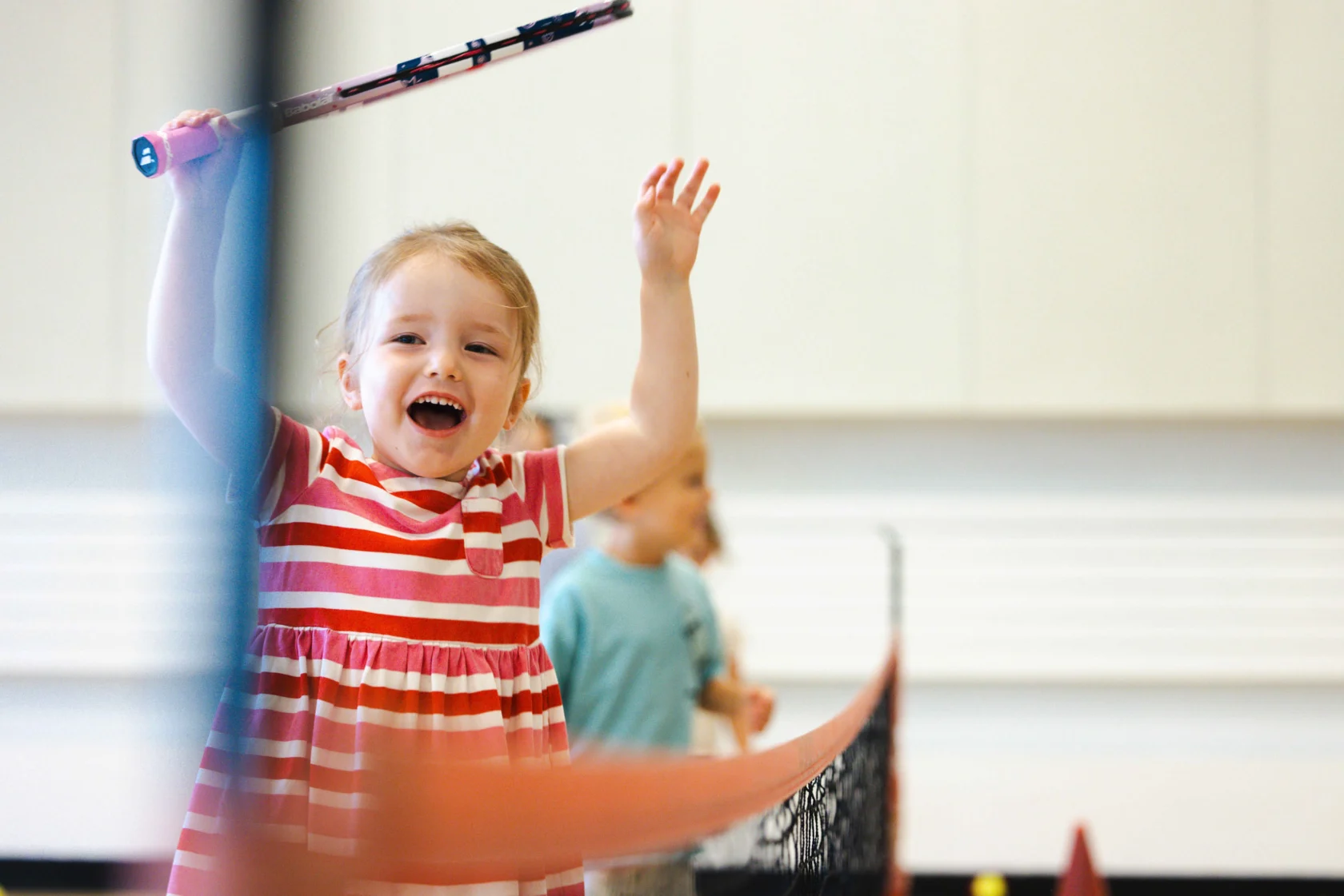 Child in a striped dress holding a tennis racket, smiling with raised arms near a net indoors.