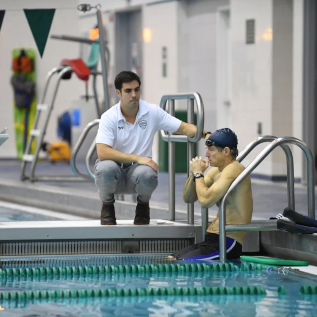 A swimmer in goggles sits at the edge of an indoor pool, talking to a coach kneeling nearby. They are next to lane dividers and a starting block.