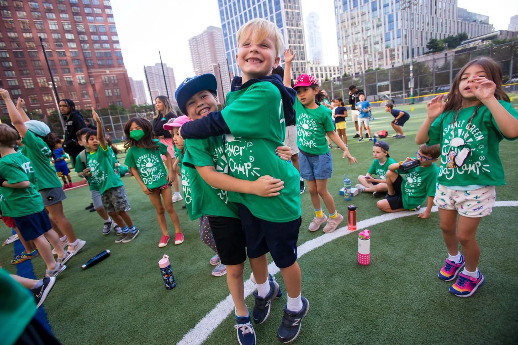 Children in green shirts play and interact on a sports field surrounded by city buildings.
