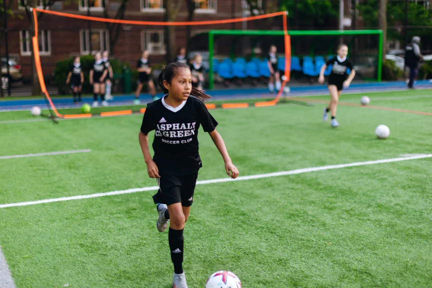 A girl in a black soccer uniform runs on a green field during practice, surrounded by soccer balls and goal nets in the background.