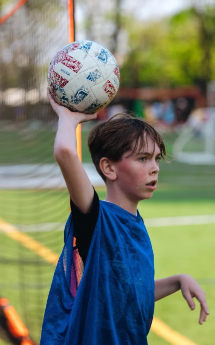 A young boy in a blue sports bib prepares to throw a soccer ball on a grassy field, with a goal net in the background.