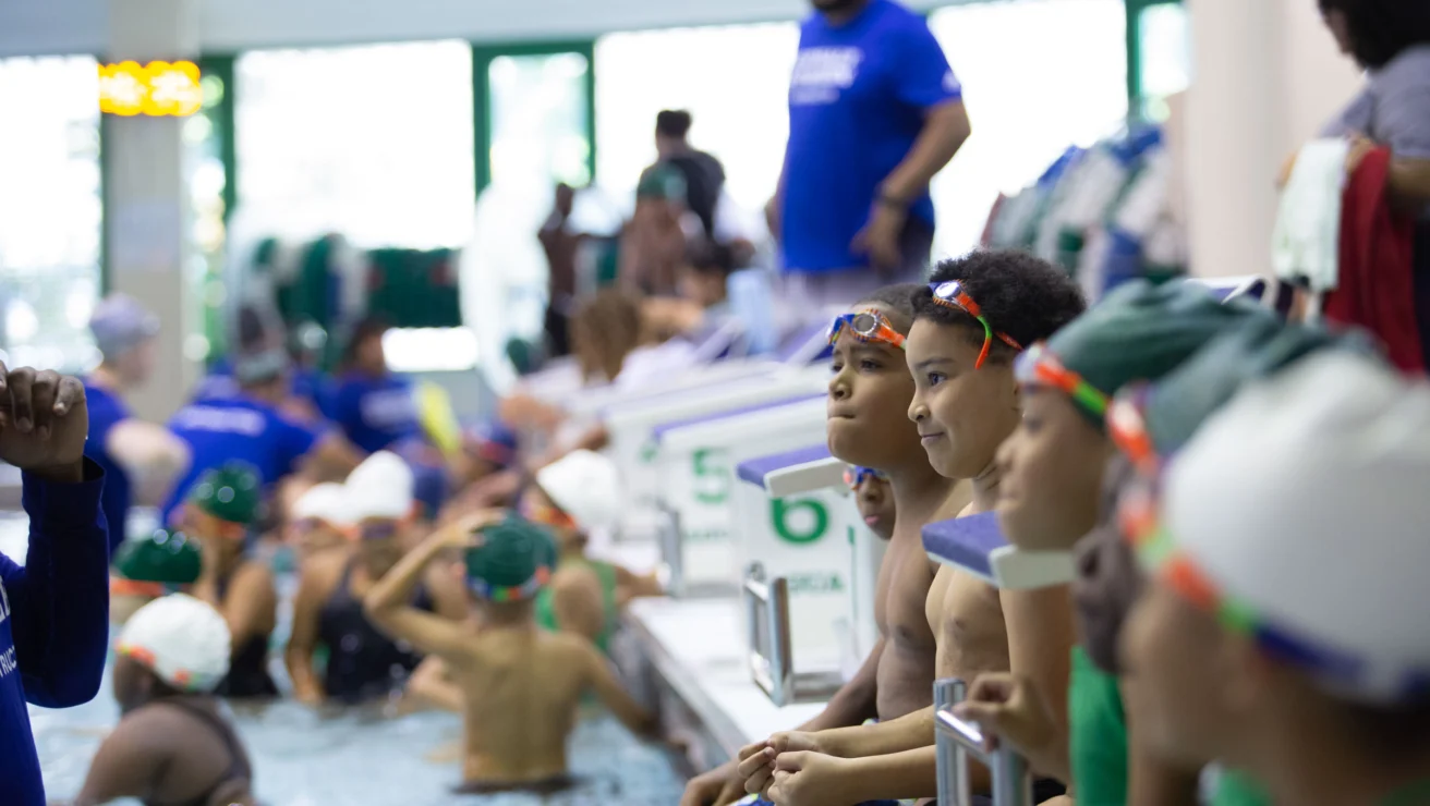 Children in swim gear sit by a pool, watching swimmers in the water. A coach in a blue shirt stands in the background.