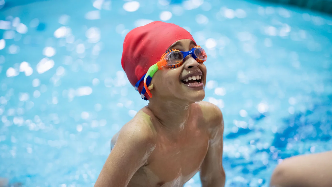 Child wearing a red swim cap and colorful goggles smiles while standing in a swimming pool.