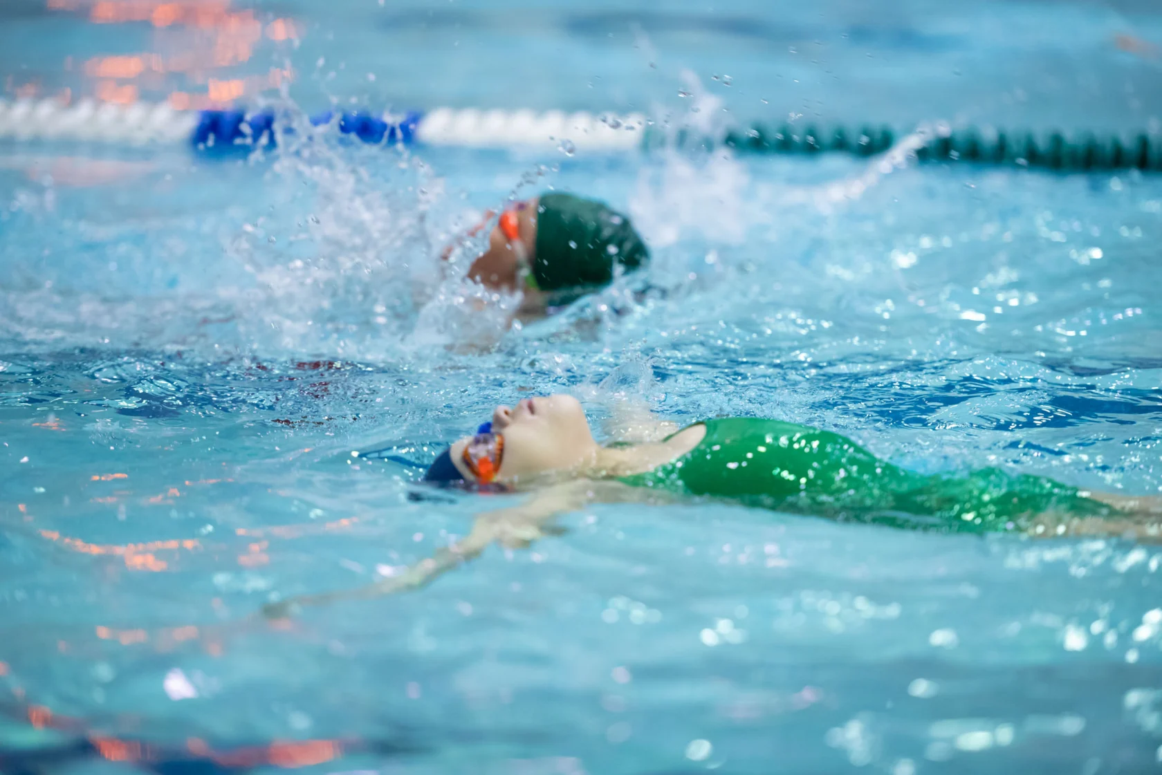 Two swimmers wearing green caps and goggles compete in a pool, creating splashes as they swim.
