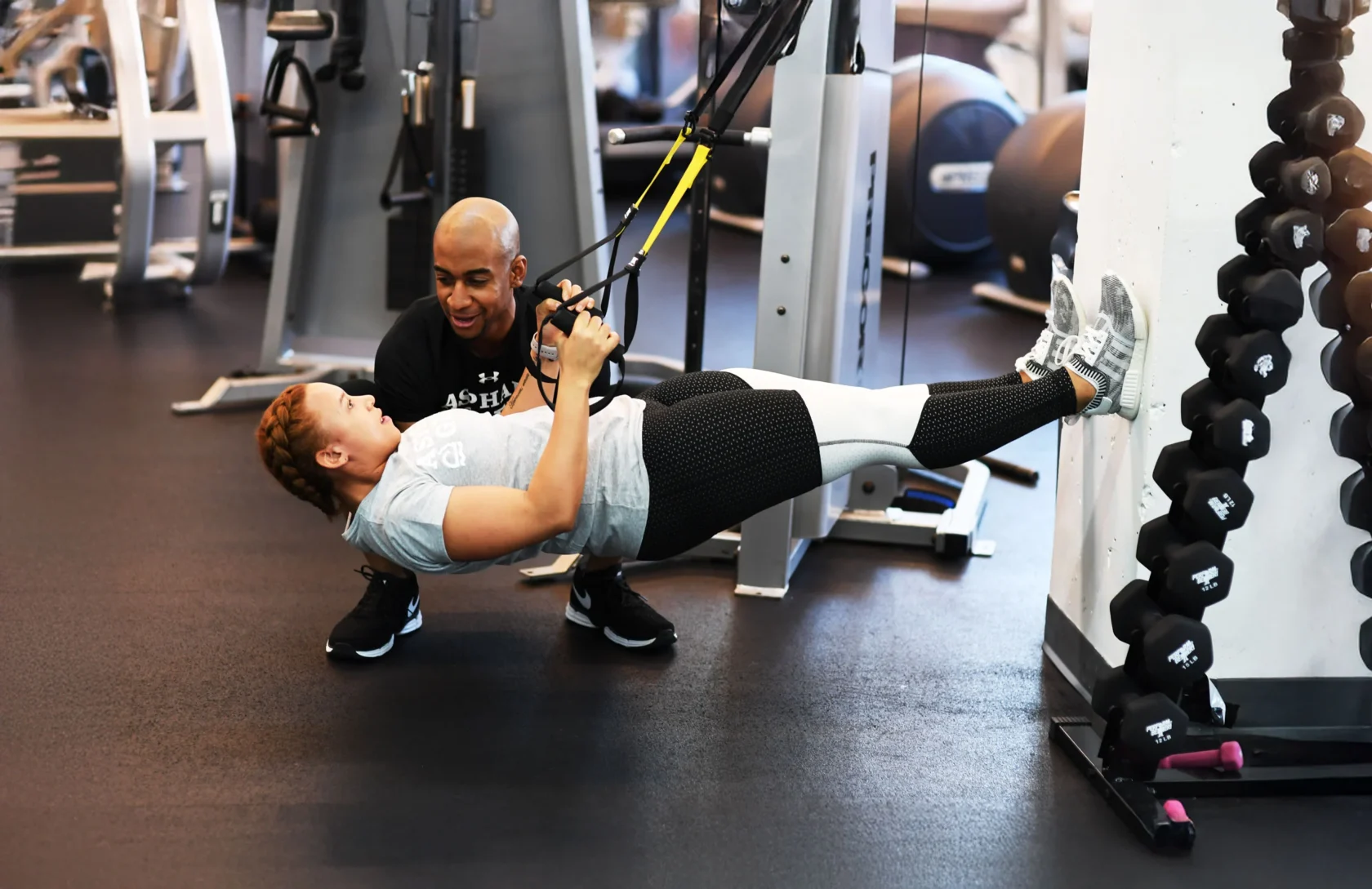 A person performs a suspension cable exercise, leaning back with feet against a wall, while another person assists in a gym setting.