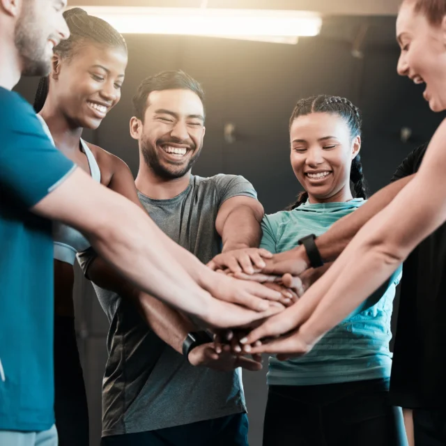 A group of people in athletic wear stand in a circle with their hands stacked together, smiling and laughing in a gym setting.
