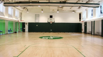 Empty indoor basketball court with wooden flooring, green padding on walls, and a hoop at the far end. Ceiling lights illuminate the space.