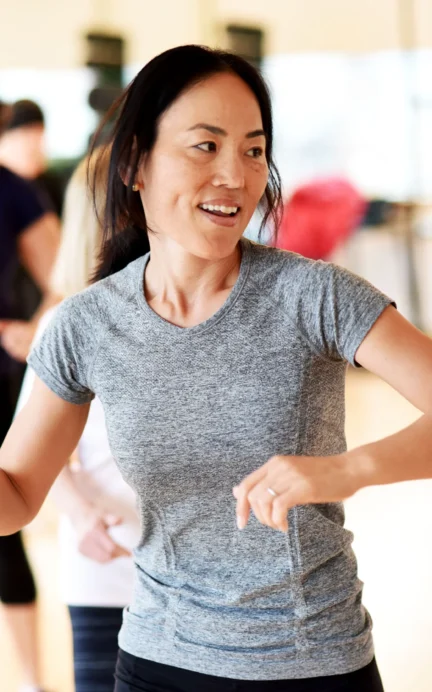 A woman in a gray shirt is smiling and participating in a group exercise class in a gym. Other participants are blurred in the background.